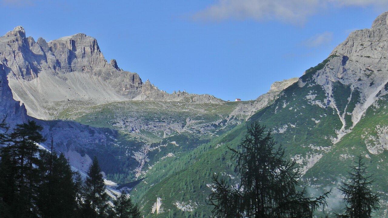 Pale limestone walls above the Fischleintal, spruce and larch on the slopes below, and the Dreizinnenhütte small on the green shoulder between them.