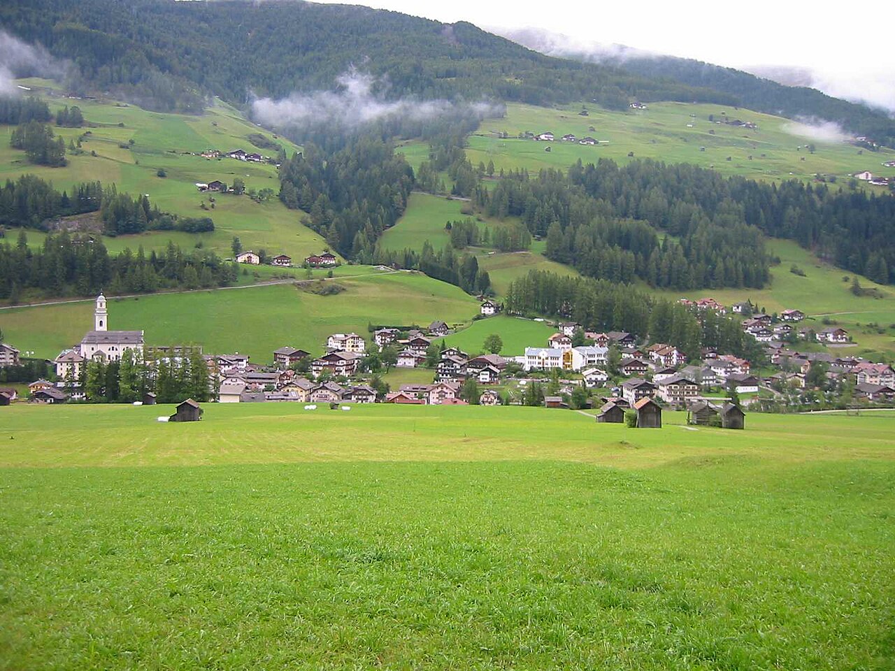 Mown meadows and hay barns below the village of Sexten, with cloud drifting across forested slopes.
