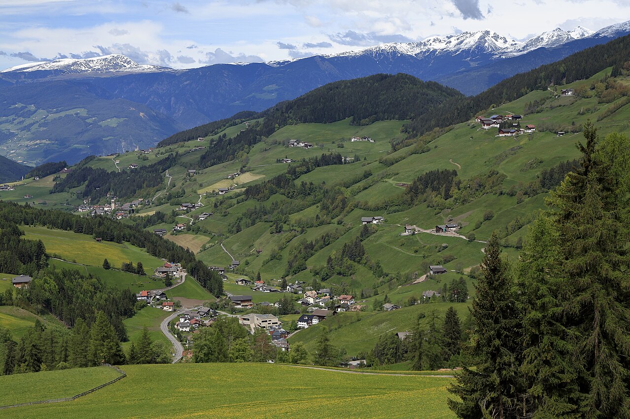 The Villnoess valley seen from the Zans road, with scattered farmsteads on green pasture and snow on the far ridge.