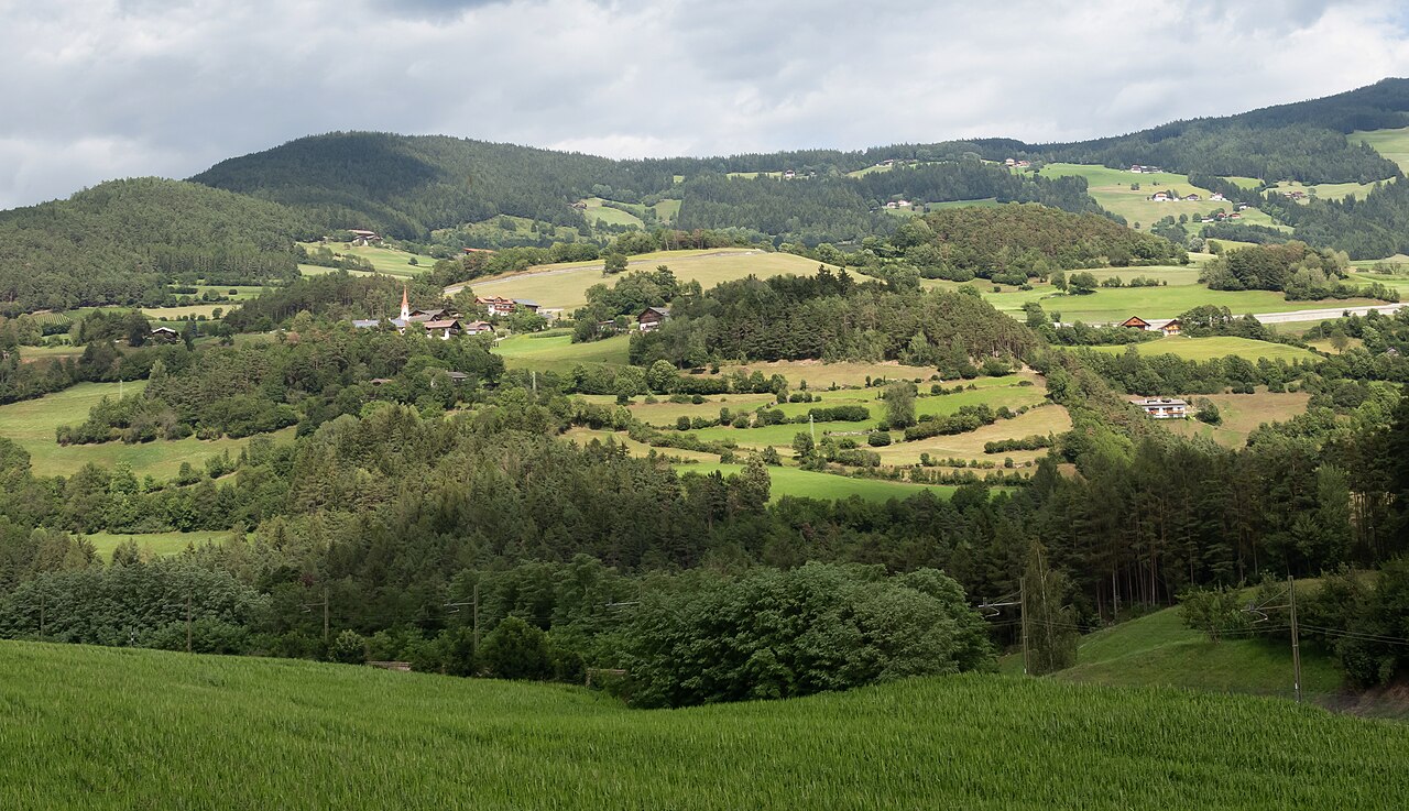 Hay meadows, mixed woodland and scattered farms on the shelf at Rodeneck, the village church spire among them, under a grey summer sky.