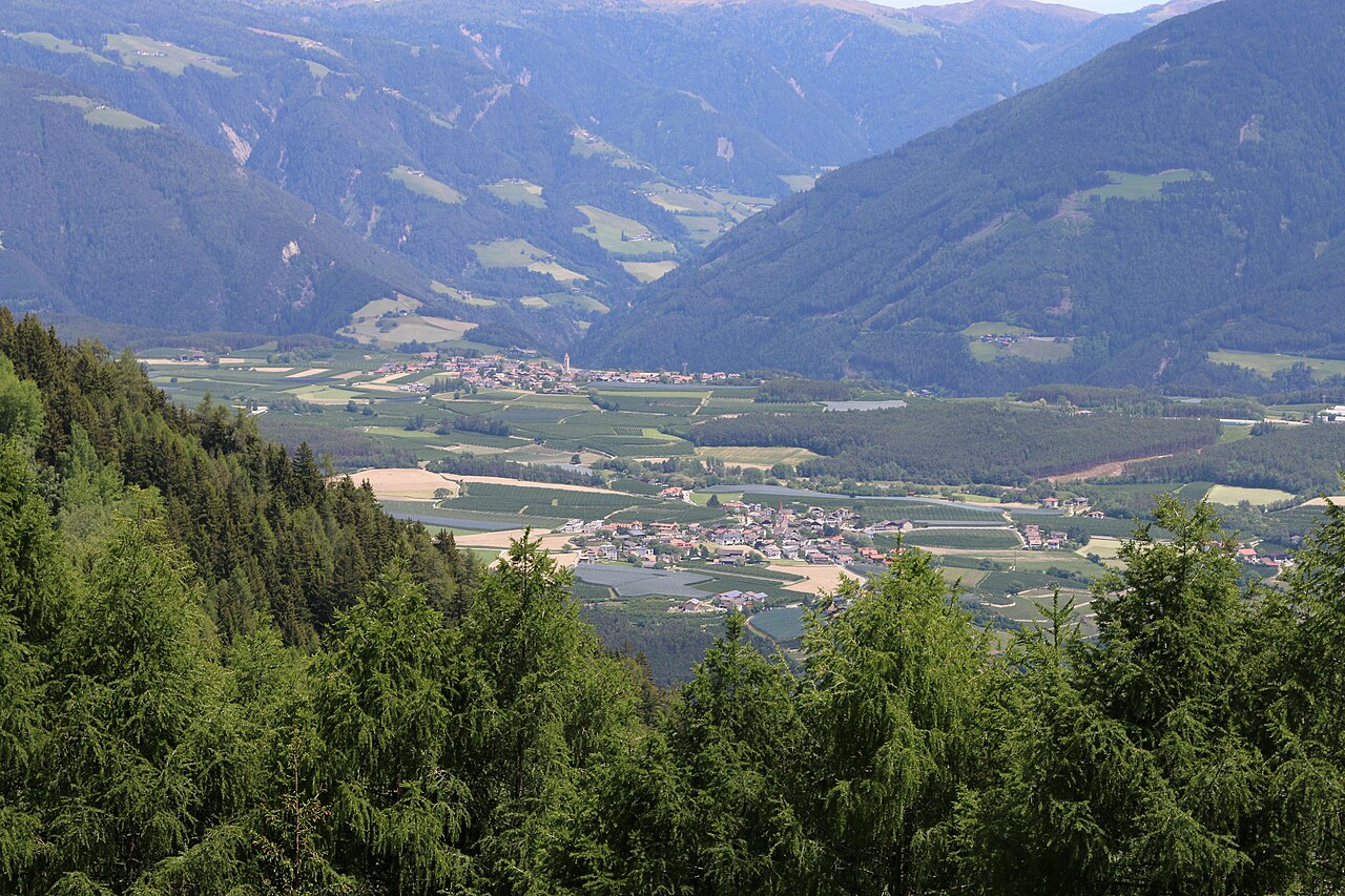 The villages of Natz and Raas among orchards and meadows on their plateau, with wooded slopes and the Luesen valley beyond.