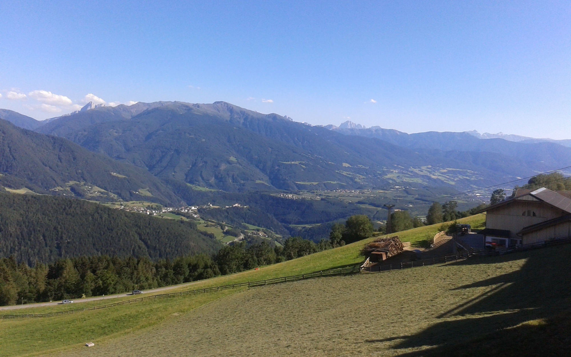 Mown pasture and timber fencing above Meransen with a farm at the right, the Eisack valley below and the Dolomites along the horizon.