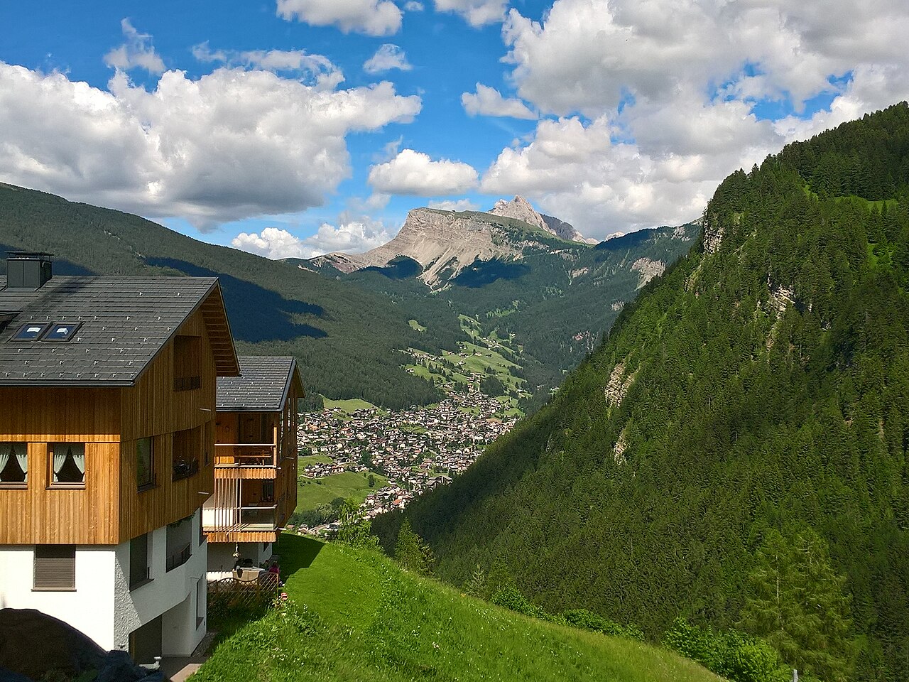 The hamlet of Bulla above Ortisei, meadow and spruce falling to the valley with the Odle wall behind, an apartment chalet at the left edge of the frame.