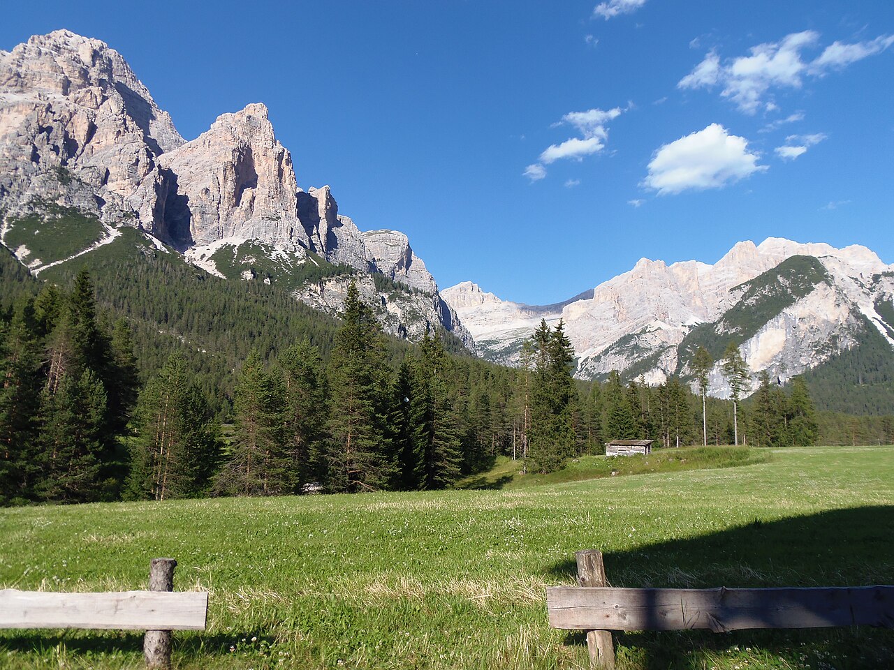 A wooden fence along the Armentara meadows, with spruce and the pale walls of the Dolomites beyond.