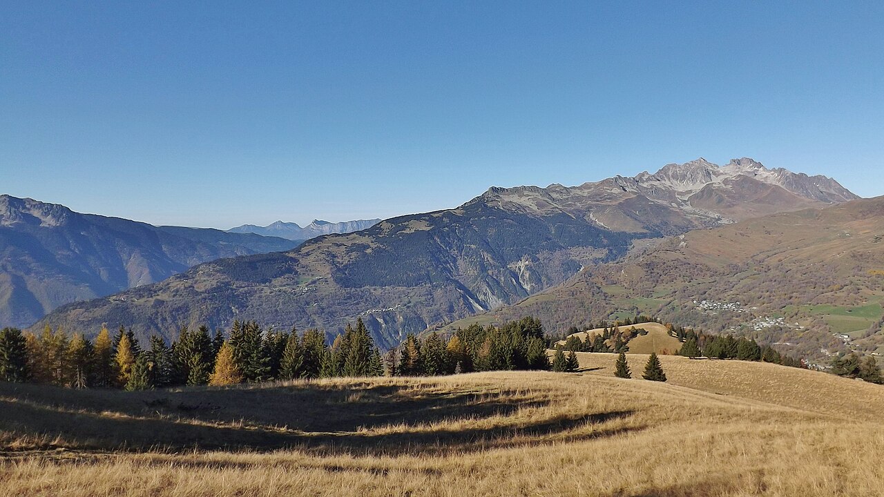 Autumn pasture and turning larch above Montpascal, with the Lauzière massif across the valley under a clear sky.