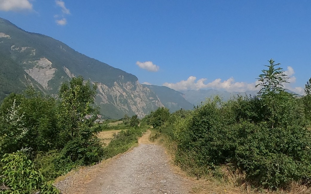 A gravel track running away between hedgerow scrub above the Maurienne with a limestone cliff face and hazy ridges beyond