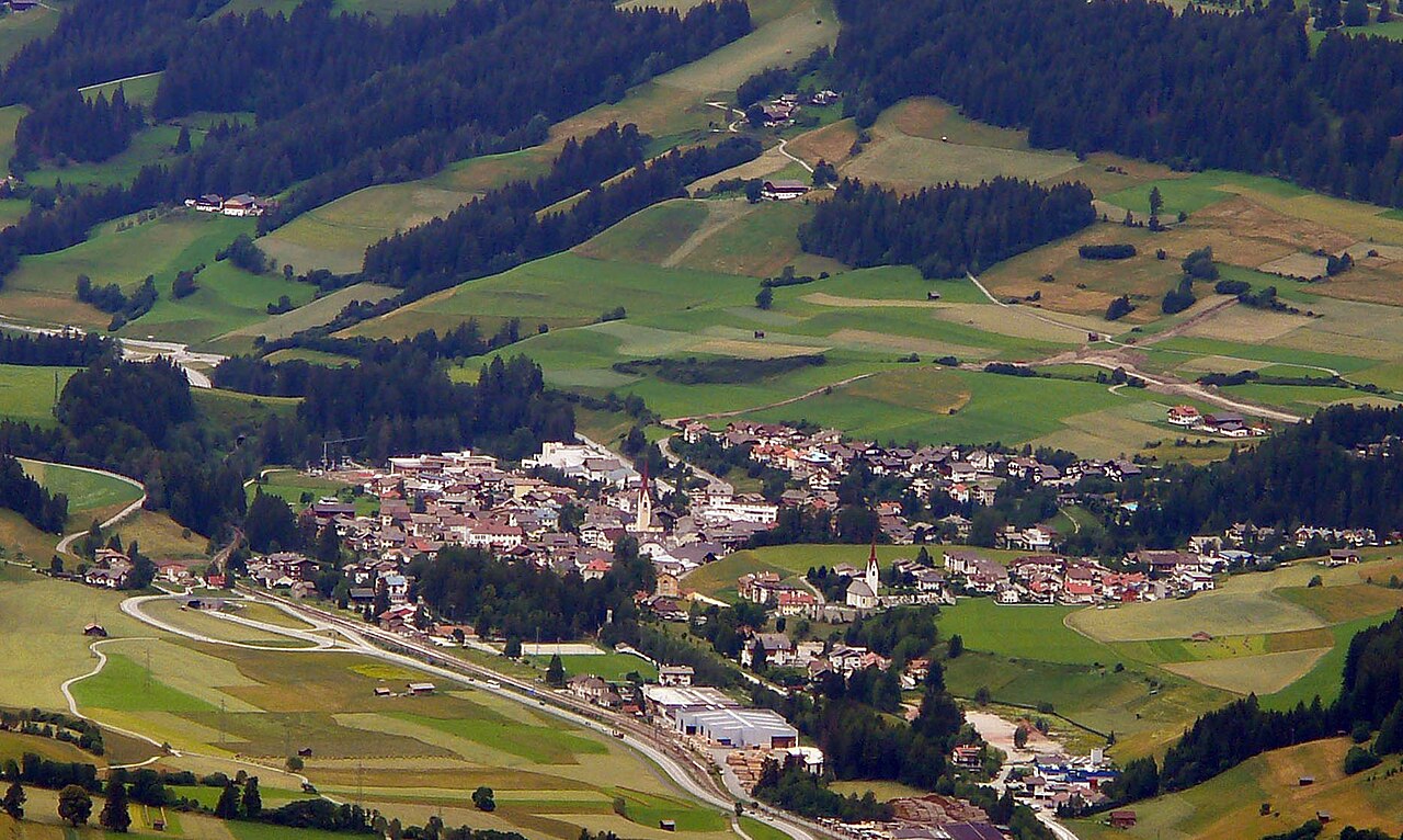 Welsberg and Tesido spread along the Puster valley floor, with mown meadows and field tracks climbing the wooded slopes above.