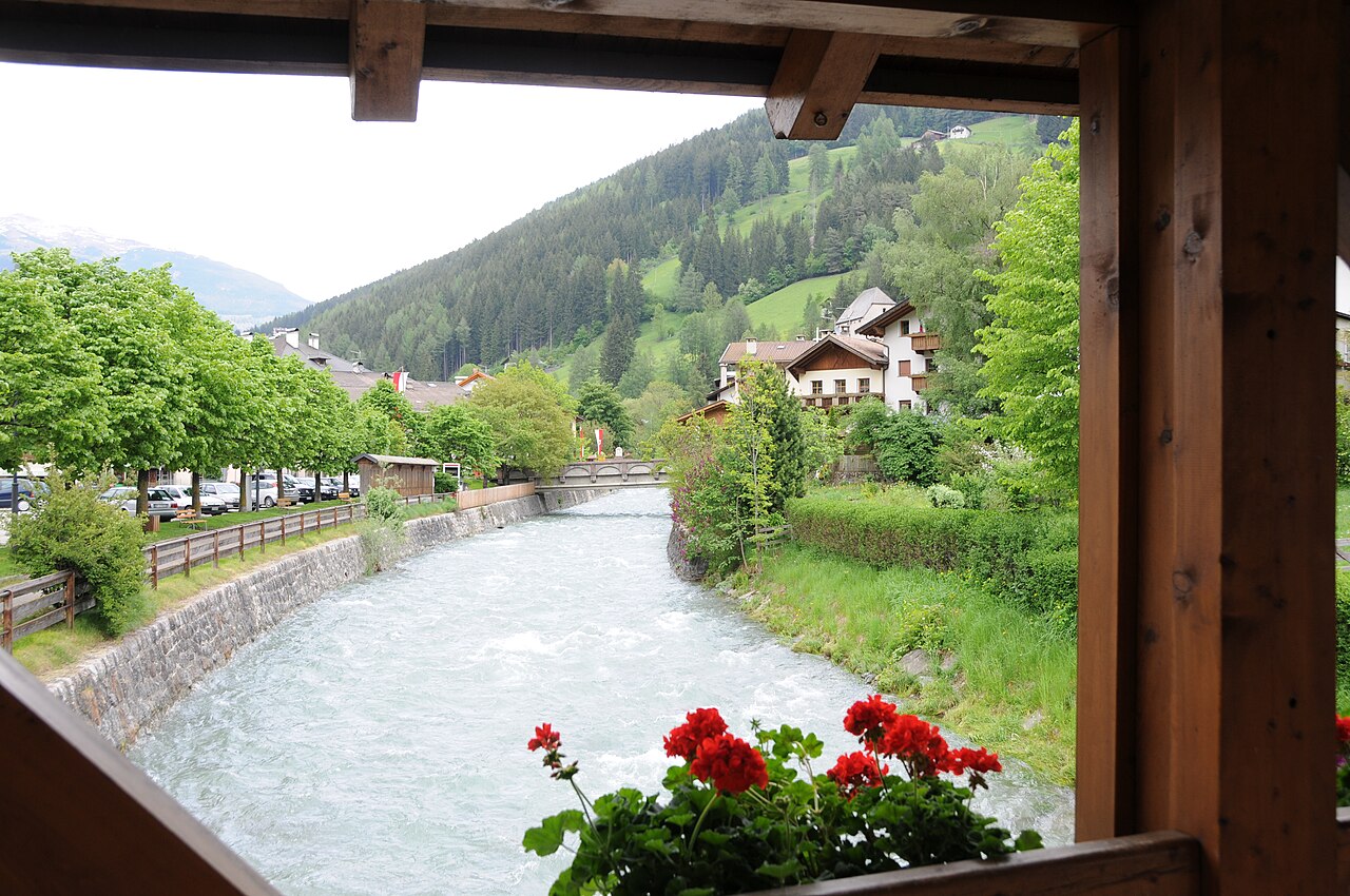 The Talfer running milky under a covered wooden bridge at Sarnthein, geraniums on the rail and the wooded valley side beyond.
