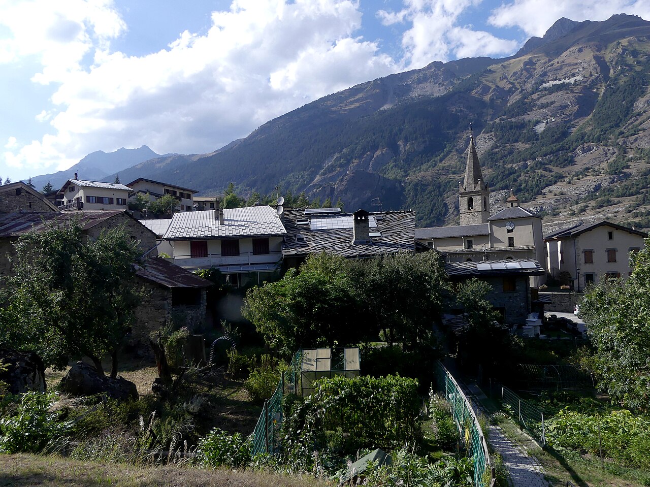 Villarodin in late afternoon, slate roofs and a church tower among kitchen gardens, a paved lane climbing between them and the Maurienne slopes behind.