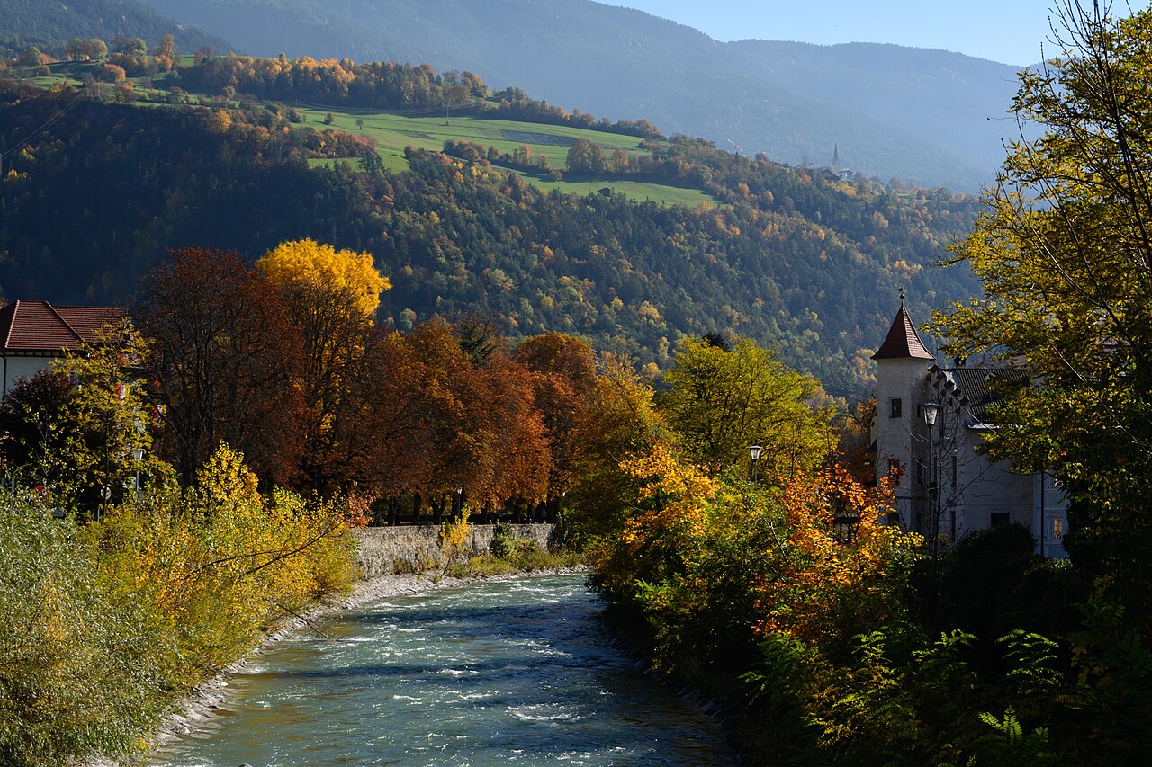 The Eisack running past a stone embankment walk at Brixen in October, beech and maple turning gold and copper along the bank, terraced meadows on the wooded valley side beyond.