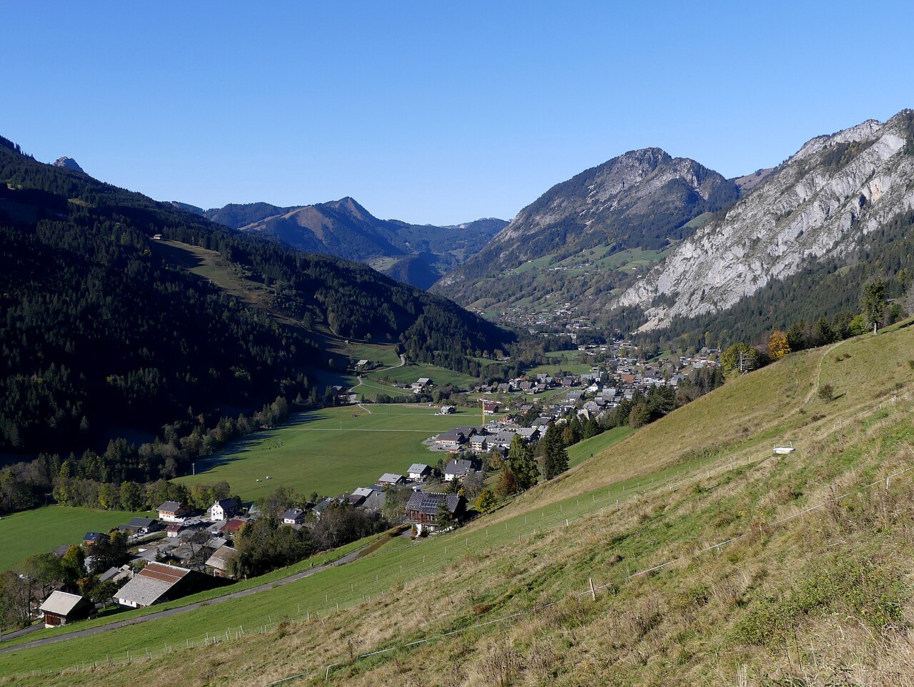 The Abondance valley on an autumn morning, its village on the floor and a path running along the grassy slope above.