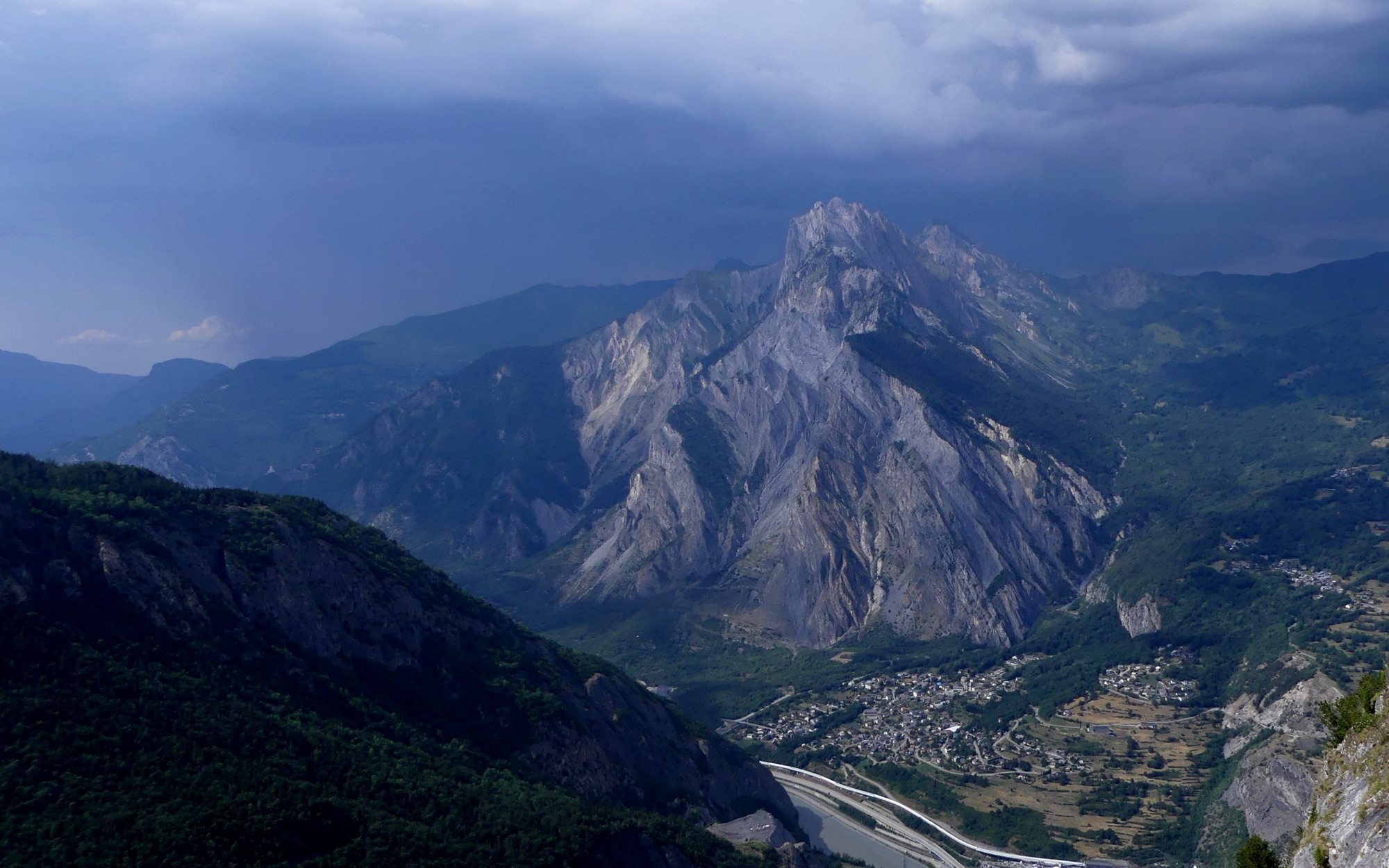 The Croix des Têtes across the Maurienne under a June storm sky, wooded crests in the foreground and the valley floor with its town far below.