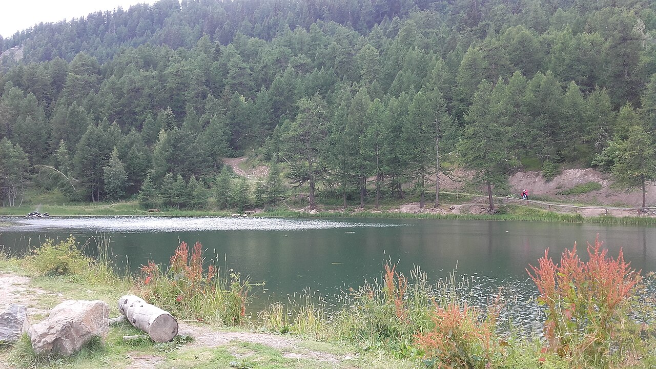 The Lac de Pramol in August, dark green under a wooded slope, with the shore path and a walker on the far bank.