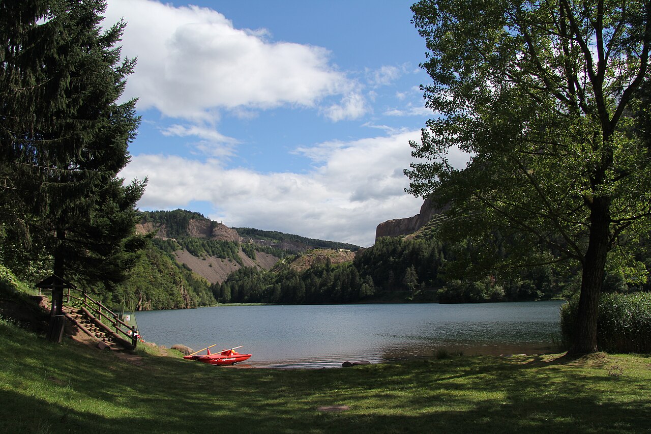 The lake at Lases seen across a grass shore under spruce and poplar, wooded slopes and pale quarry faces on the far side.