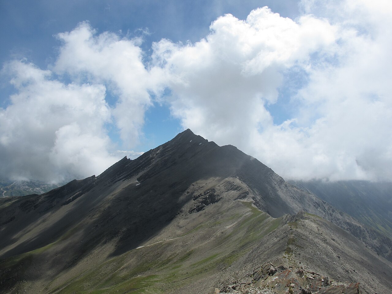 The dark schist ridge of the Grande Chible above Montricher-Albanne, a path traced along its alpine grass shoulder under building cloud.