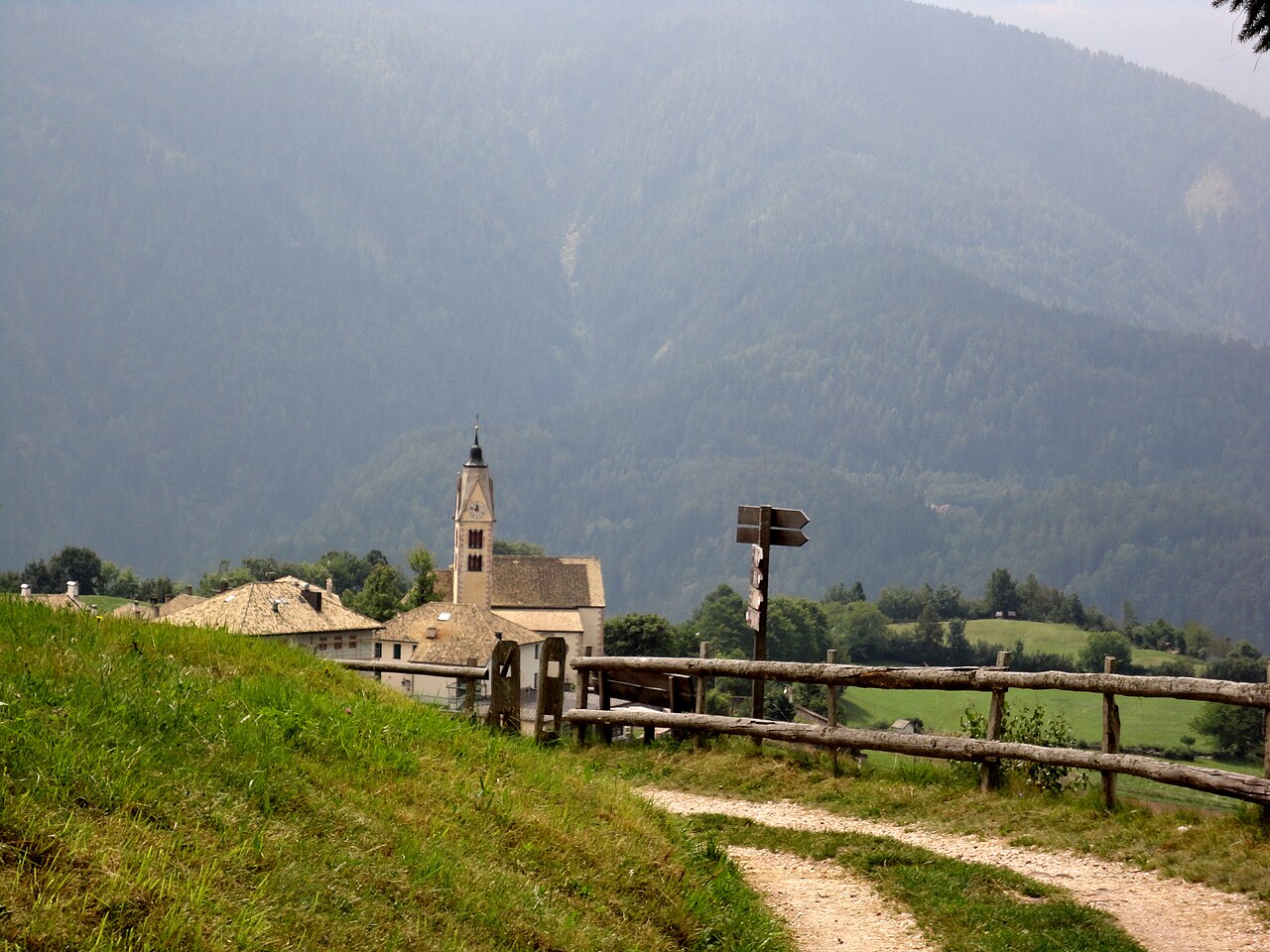 A gravel track behind a timber rail fence running towards Anterivo and its church tower, forested slopes hazy behind.