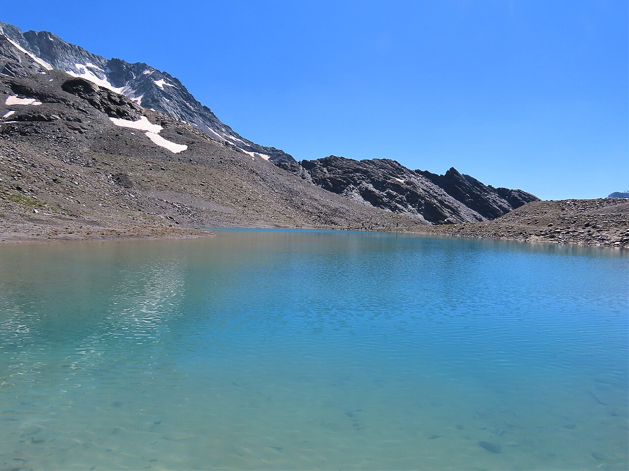 The turquoise water of the Lac Blanc above Val Thorens, clear to the stones, with summer snow patches on the peak above.