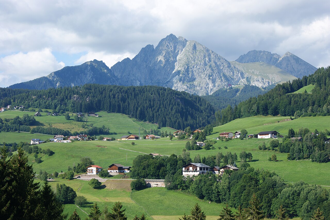 Green meadows and scattered farmhouses on the Voeran terrace, with the rock summit of the Ifinger rising behind the forest.