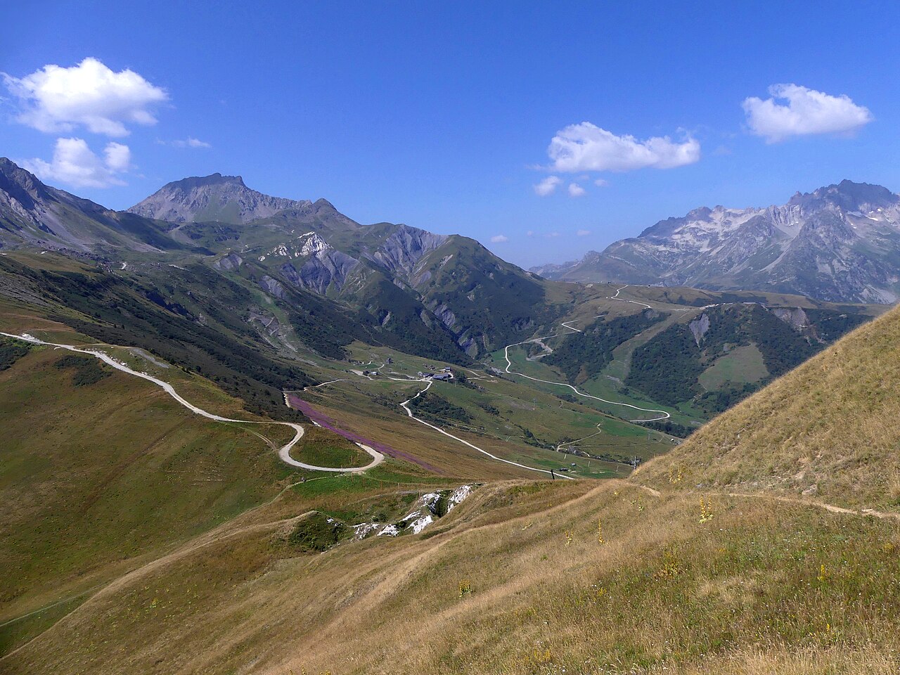 High summer pasture above Valmorel with a track winding down towards the Col du Gollet and rock peaks along the skyline.