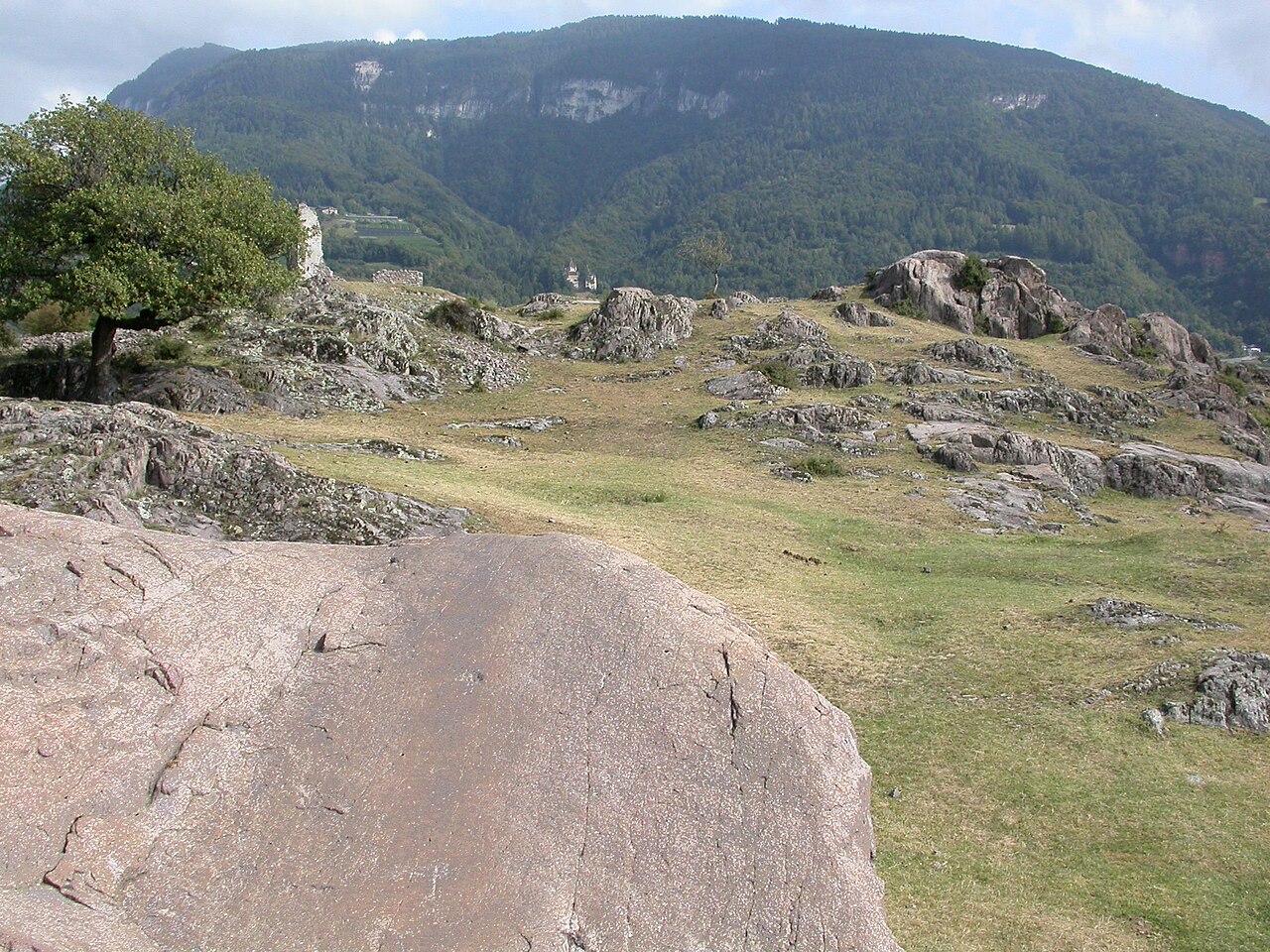 The smooth Heidenrutsche rock and dry grass on the Castelfeder hill, with ruin walls above and wooded slopes beyond.