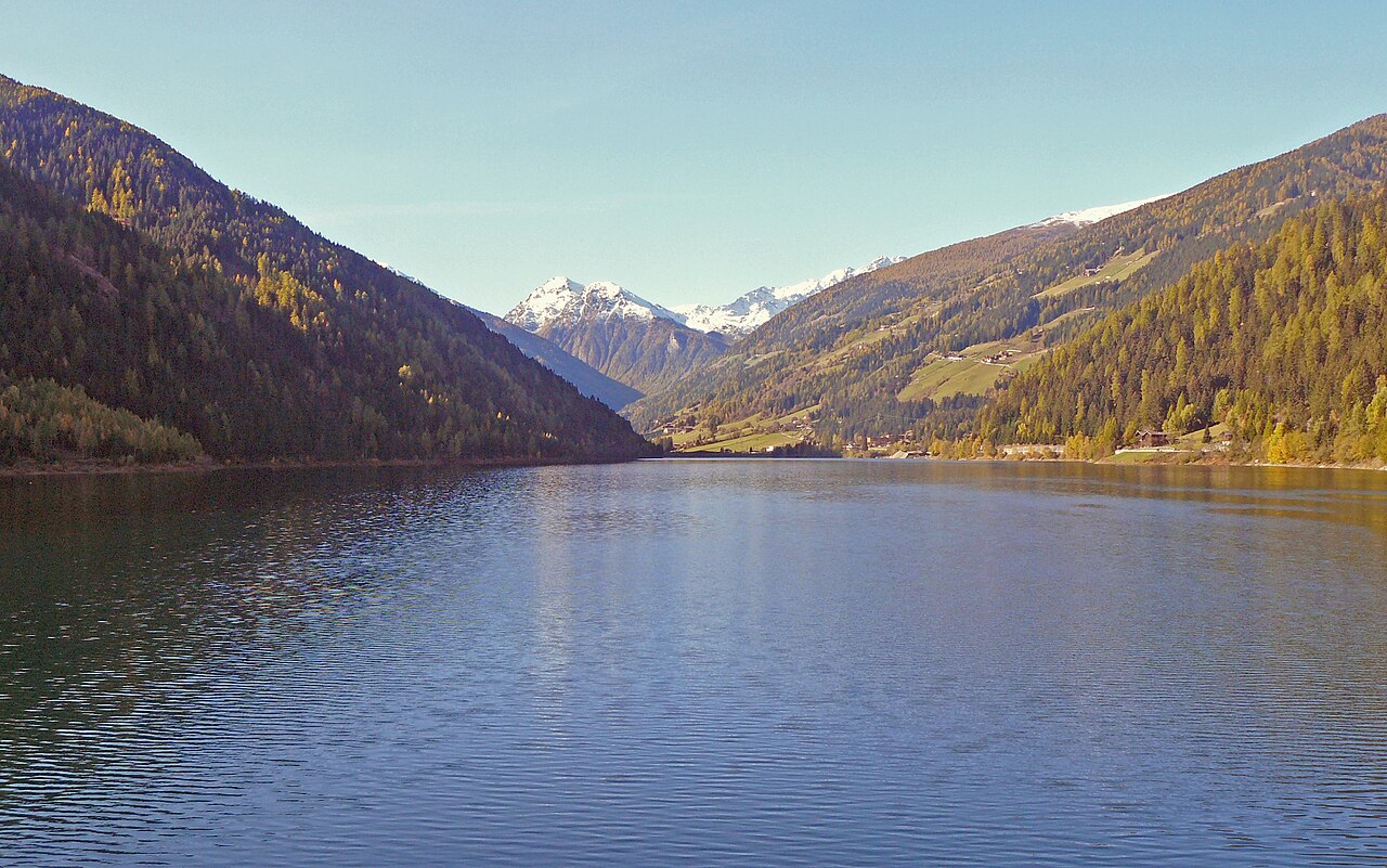 The Zoggler reservoir in the Ultental in autumn, larch turning gold on both slopes and snow on the peaks at the head of the valley.