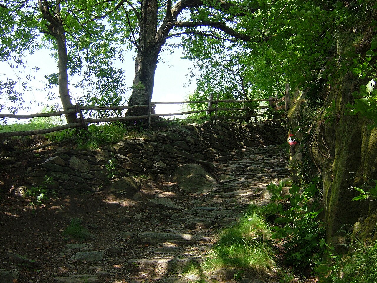A stony sunken path above Naturns between a dry-stone wall and a rustic timber fence, chestnut trees overhead and a red-and-white waymark on the rock.