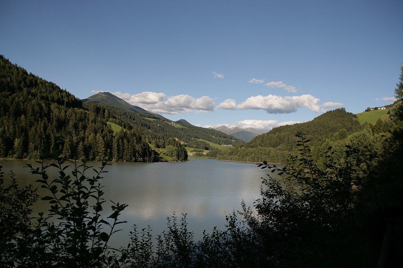The Olang lake held between wooded slopes, meadows and farms above the far shore and distant peaks beyond.