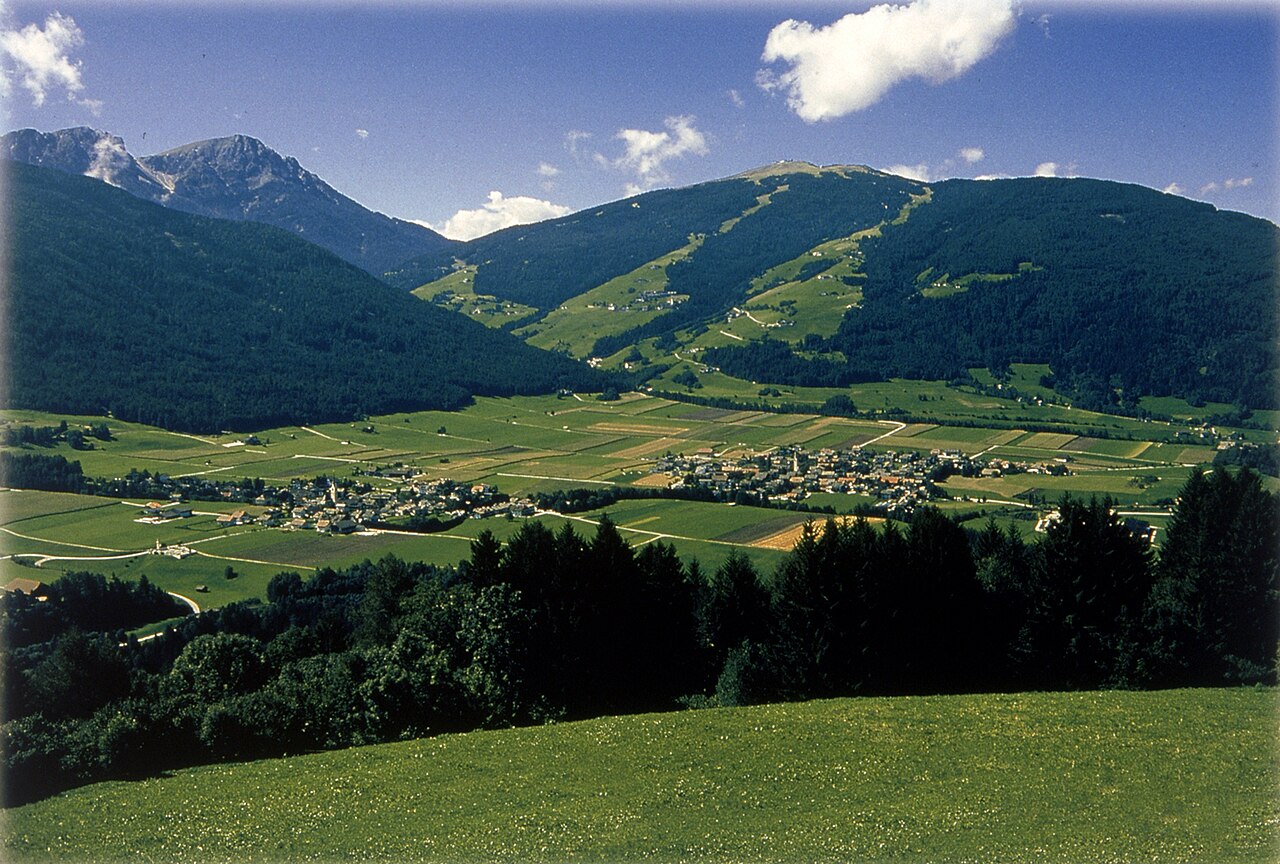 Fields and villages on the floor of the Puster valley at Olang, below wooded slopes and a rocky summit.