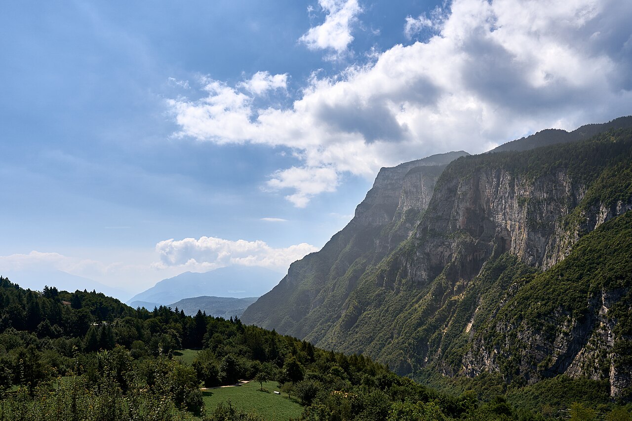 A meadow and track on the wooded terrace at Fai della Paganella, the limestone escarpment of the Paganella rising to the right under cumulus.