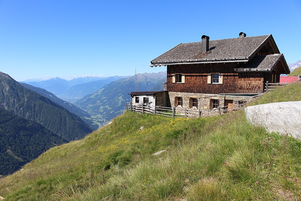 The shingled Fleckner hut on flowering alpine grass at 2100 m, with the Passeier valley falling away behind it.