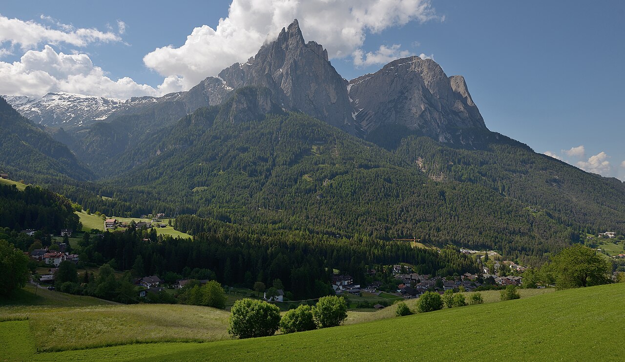 Hay meadows above Seis am Schlern with the village and forested slopes below, the Schlern and the Santner Spitze rising behind under cumulus.