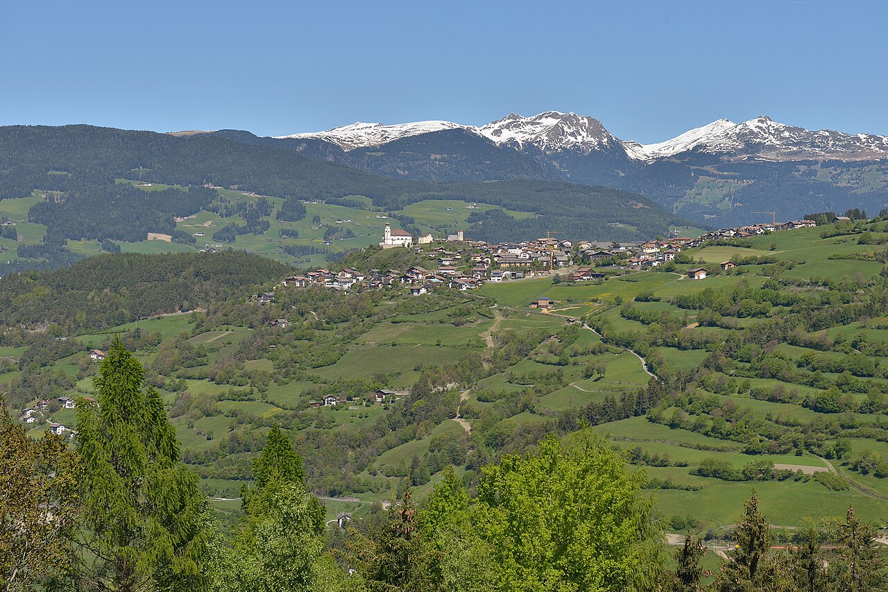 The village of Lajen on its terrace above green meadows crossed by field paths, with snow on the far range.