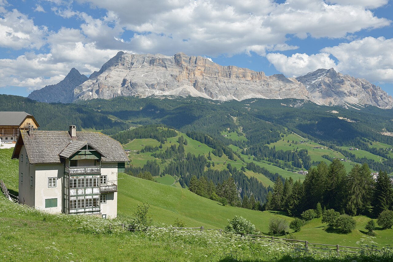 A flowering hay meadow at Pescol with an old Ladin farmhouse and timber fences, green terraces and scattered farms beyond, and the wall of the Sas dla Crusc behind.