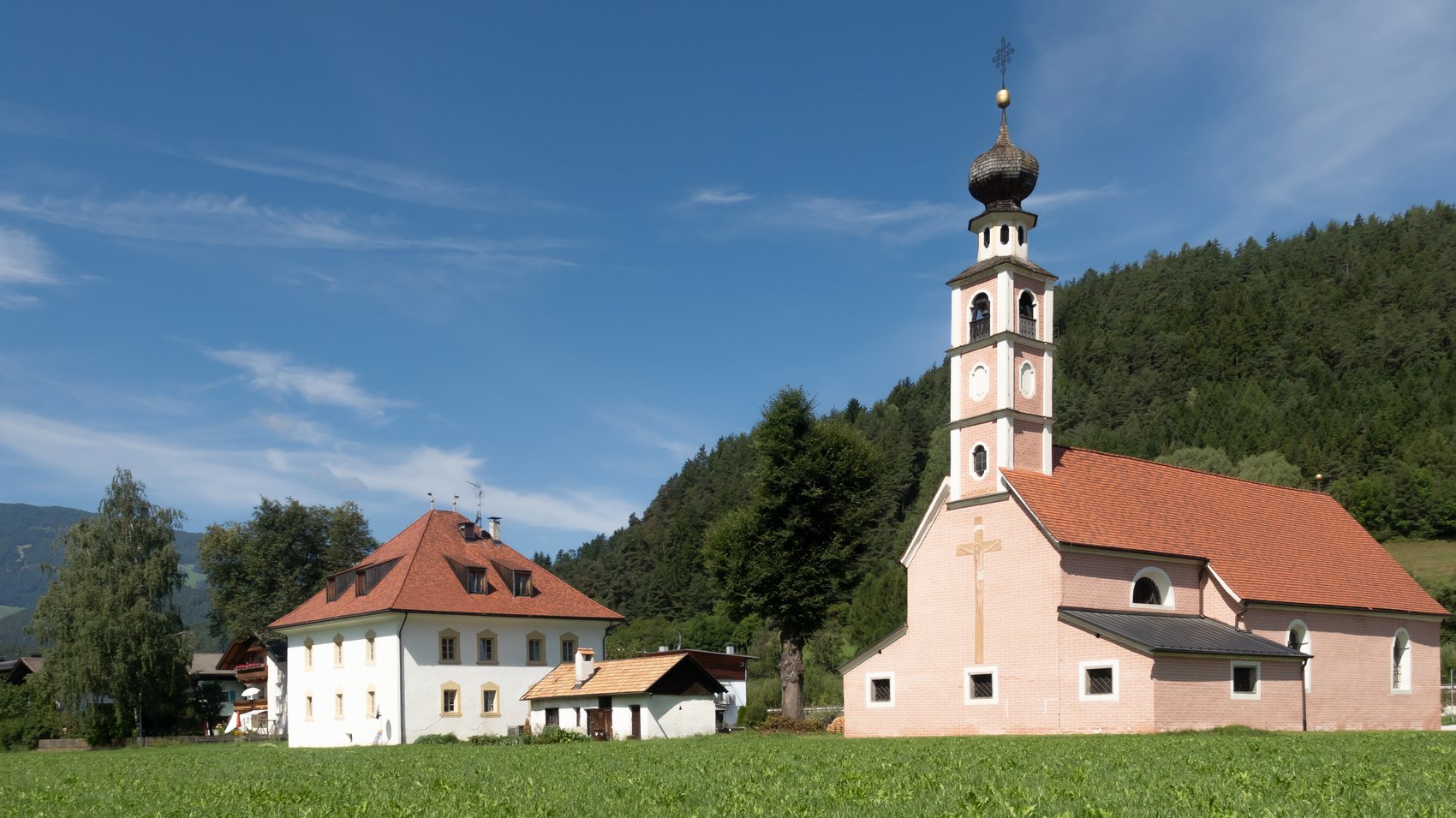 The pink church of Heiligkreuz in Fronwies and its Widum house standing on mown meadow below a wooded hill near St. Lorenzen