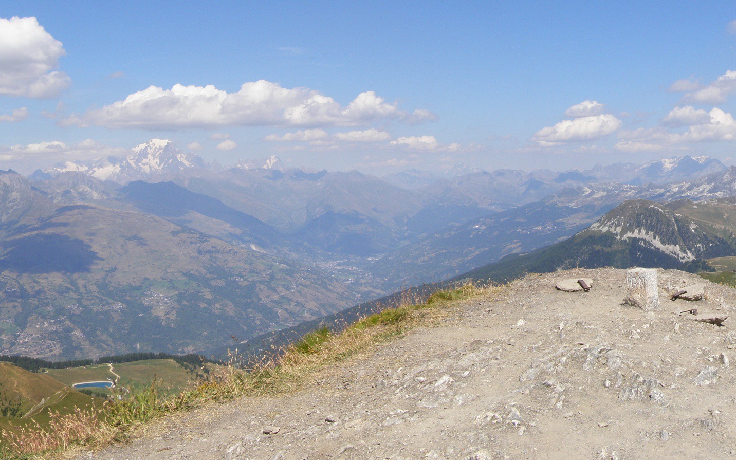 The stony summit of the Mont Jovet with its survey marker, dry grass on the shoulder and the Tarentaise valley below, Mont Blanc on the horizon.